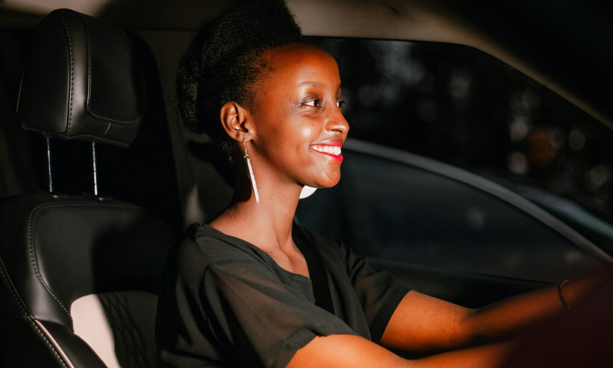 A woman with black hair smiles while driving a car during sunset.